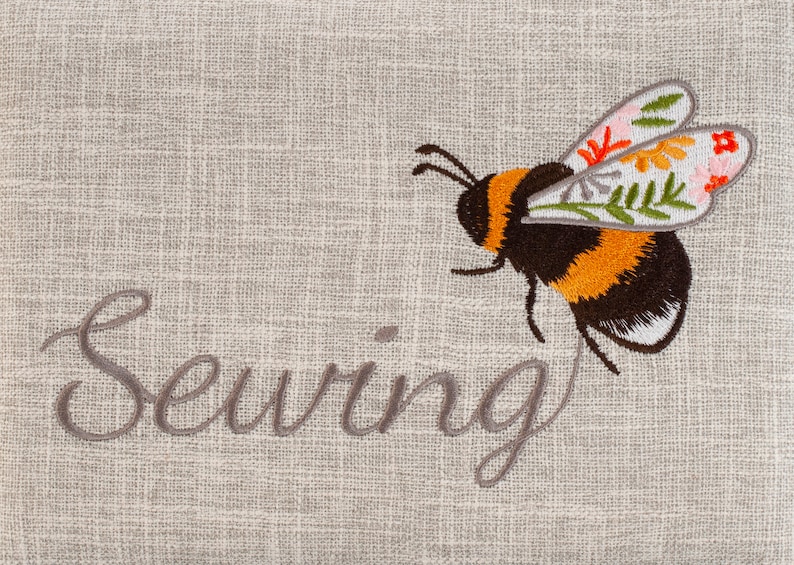 Puede incluir: Una tela de lino beige con la palabra "Costura" bordada en hilo gris. Una abeja bordada estilizada con rayas negras, amarillas y blancas vuela sobre la palabra. La abeja tiene alas florales con acentos rosas, rojos, amarillos y verdes.