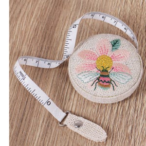 May include: A beige, round measuring tape with a floral and bee embroidery design. The tape extends to 152 cm and is white with black markings. The tape measure is made of fabric and has a metal clasp.