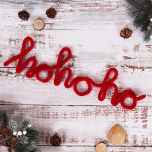 Könnte beinhalten: Weihnachtsdekoration mit dem Wort "hohoho" aus leuchtend rotem Garn. Der Text ist auf einer rustikalen weißen Holzoberfläche angeordnet, verziert mit Tannenzapfen, Holzscheiben und Tannenzweigen, die ein saisonales Urlaubsgefühl erzeugen.