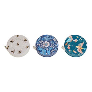 May include: Three decorative tape measures with unique designs. One features bees on a light gray background, another showcases a blue floral pattern, and the third has birds and flowers on a teal background. Each tape measure has a silver metal pull.