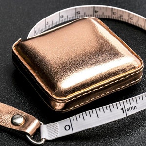 May include: A rose gold measuring tape with a square case and a white tape marked in inches. The tape is 60 inches long.