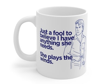 May include: White ceramic mug with a blue illustration and text. The mug features a drawing of a man and the quote, "Just a fool to believe I have anything she needs. She plays the winds."