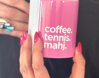 May include: A two-tone ceramic mug held in hands. One side is white, the other is bright pink with the words "coffee. tennis. mahj." printed in white. The mug is cylindrical and appears to be empty.