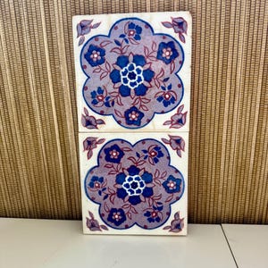 May include: Two square ceramic tiles with a floral design. The tiles feature a lavender background with blue and white flowers. The flowers are arranged in a symmetrical pattern. The tiles are on a white surface.