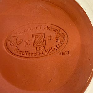 May include: A terracotta-coloured ceramic plate base with an embossed oval logo. The logo reads "Victoria and Richard", "M", "1989", and "MacKenzie-Childs, Ltd." with decorative elements. The year "1983" is also embossed.