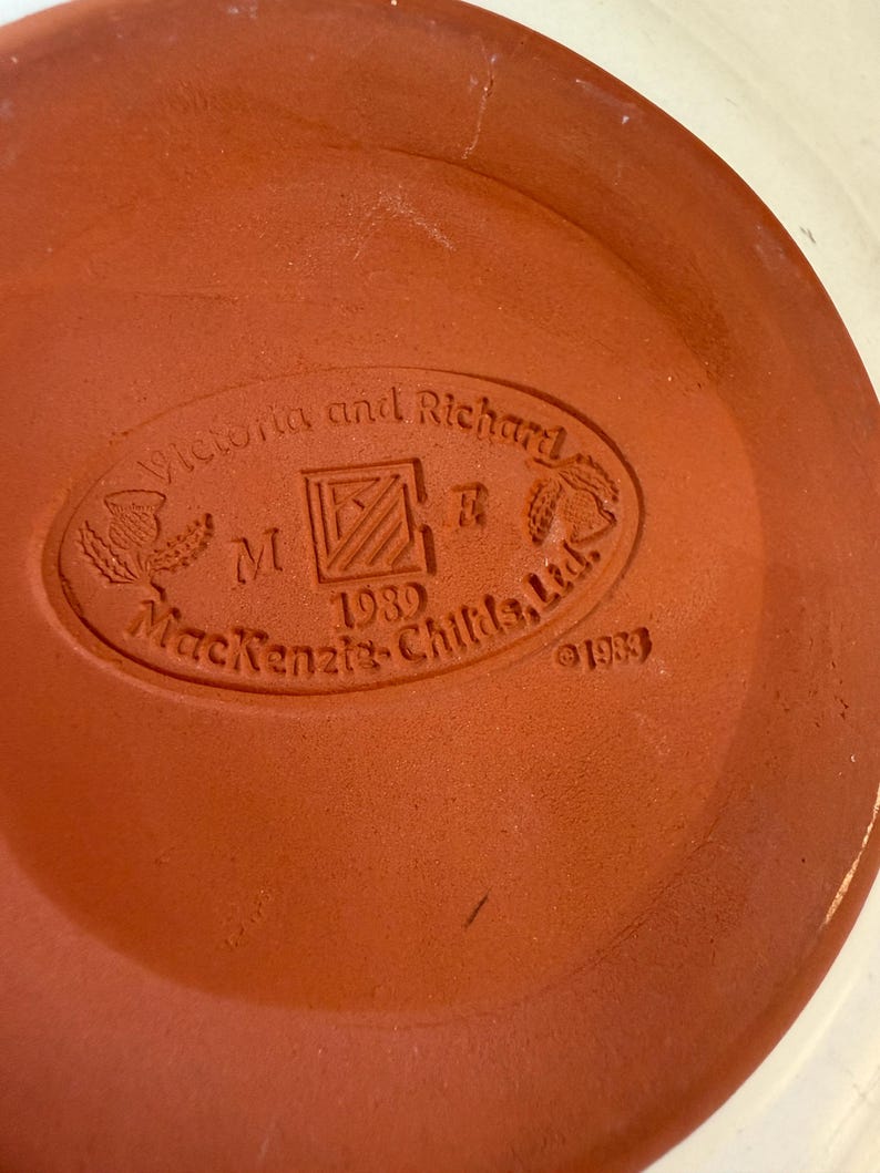 May include: A terracotta-coloured ceramic plate base with an embossed oval logo. The logo reads "Victoria and Richard", "M", "1989", and "MacKenzie-Childs, Ltd." with decorative elements. The year "1983" is also embossed.