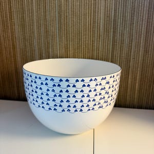 Puede incluir: Cuenco de cerámica blanco con un patrón de tréboles azules alrededor del borde. El cuenco tiene forma redondeada y un acabado liso y brillante. El fondo es una superficie texturizada de color marrón claro.