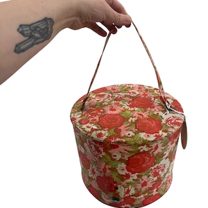 Könnte beinhalten: Eine runde Hutbox mit floralem Muster und einem einzigen Henkel. Die Box ist rot, grün und weiß mit einem floralen Muster.