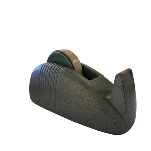 May include: A heavy, dark grey cast iron tape dispenser with a textured, matte finish. The dispenser features a rounded, streamlined body with vertical ribbed detailing on the side and a serrated metal cutting edge at the front for tape.