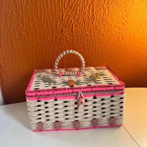 May include: A small woven wicker box with a pink and black checkered pattern. The box has a pink trim and a wooden handle. The lid is decorated with embroidered flowers.