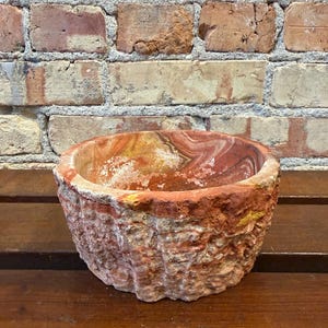 May include: A round stone bowl featuring a rough, textured exterior and a smooth, polished interior. The material displays natural swirling patterns in shades of reddish-brown, orange, and cream, with a thick, uneven rim and a sturdy, heavy base.