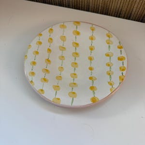 May include: A circular, white ceramic plate with a pink edge. The plate features a repeating pattern of yellow floral shapes linked by green stems. The design is evenly spaced across the plate's surface.