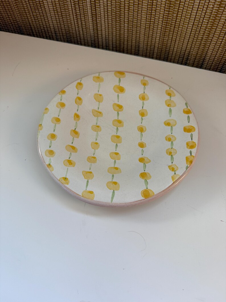 May include: A circular, white ceramic plate with a pink edge. The plate features a repeating pattern of yellow floral shapes linked by green stems. The design is evenly spaced across the plate's surface.