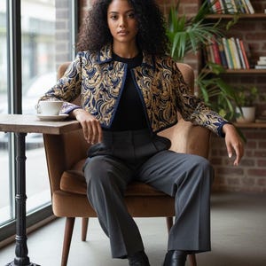 Puede incluir: Una persona sentada en una silla, vistiendo una chaqueta estampada en oro y azul marino, una blusa negra y pantalones grises. Lleva botas negras. Una pequeña mesa con una taza y un platillo está a su izquierda. El fondo incluye una ventana y una pared de ladrillos.