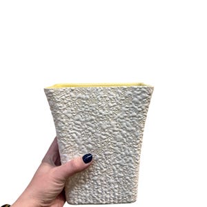 May include: Cream-colored, textured ceramic vase with a square shape and a yellow interior. The vase has a rough, bumpy surface and a slightly flared design. A hand is holding the vase.