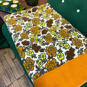 May include: A vintage floral blanket with a white background and large orange, yellow, and brown flowers. The blanket has an orange border and is draped over a green chair. The blanket's design is reminiscent of the 1970s.