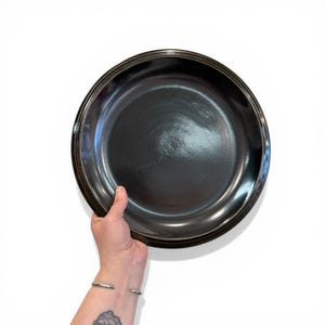 May include: A black, round serving dish with a glossy finish. The dish has a slightly raised rim and is held by a person's hand. The dish is likely made of ceramic or a similar material and is suitable for serving food.