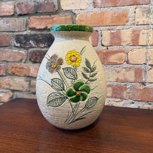 May include: A cream-coloured ceramic vase with a textured surface. The vase features hand-painted flowers in yellow, brown, and green, with green leaves and stems. The rim is painted green. The vase is on a wooden surface.