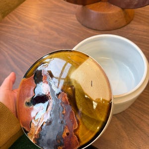 May include: A ceramic lidded bowl with a glossy, abstract design on the lid. The lid features a blend of brown, gold, and black hues. The bowl itself is a light cream color. The bowl is open, revealing its interior.