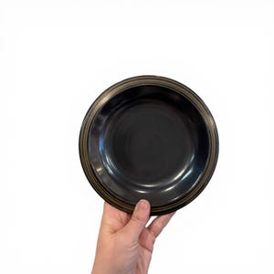 May include: A black ceramic bowl with a gold rim is held in a hand against a white background. The bowl has a simple, elegant design, suitable for serving food. The bowl's interior is a glossy black.