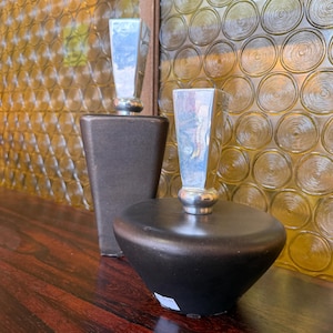 May include: Two decorative perfume bottles with silver stoppers. One bottle is a dark brown, rectangular shape, and the other is a black, rounded shape. The background features a textured, yellow-tinted glass window.