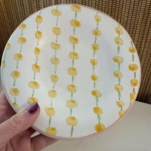 May include: White ceramic plate featuring a hand-painted design of yellow floral shapes on green stems. The plate has a pink rim. The plate is held by a hand with purple nail polish.