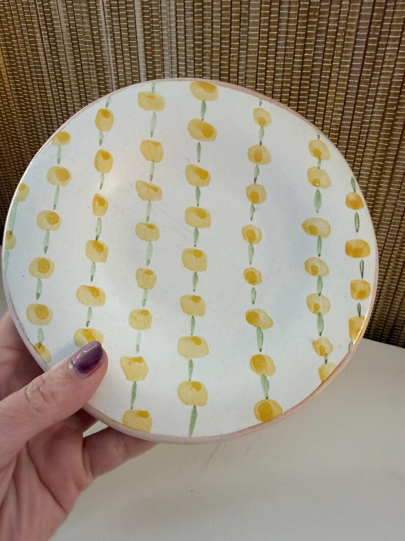 May include: White ceramic plate featuring a hand-painted design of yellow floral shapes on green stems. The plate has a pink rim. The plate is held by a hand with purple nail polish.