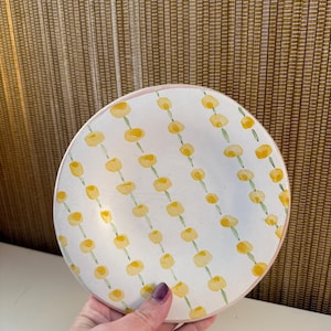 May include: A round, white ceramic plate with a pink rim. The plate is decorated with a repeating pattern of yellow floral designs. The plate is held by a person.
