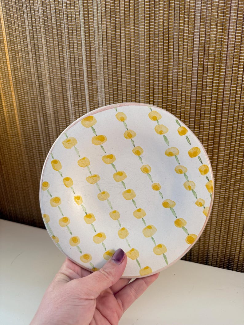 May include: A round, white ceramic plate with a pink rim. The plate is decorated with a repeating pattern of yellow floral designs. The plate is held by a person.