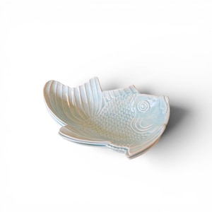 Puede incluir: Un plato de cerámica azul claro con forma de pez. El plato presenta escamas, aletas y cola detalladas. El plato se utiliza probablemente para servir aperitivos o porciones pequeñas. El plato está sobre un fondo blanco.