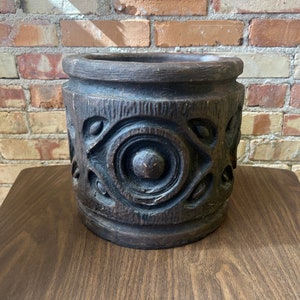 Può includere: Un vaso di legno marrone scuro con un design intagliato. Il vaso ha un design circolare al centro con un pomo in rilievo.