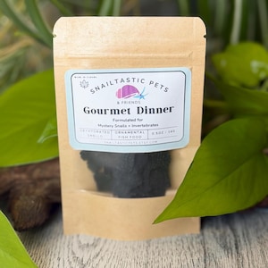 May include: A brown paper pouch containing dark, dehydrated food for snails and invertebrates. The label reads Snailtastic Pets Gourmet Dinner, Formulated for Mystery Snails + Invertebrates, Dehydrated Snello, Ornamental Fish Food, 14g / 0.5oz.