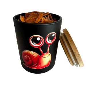 May include: Black ceramic container with a red snail graphic and a bamboo lid. The snail has large eyes and a smiling face. The container is filled with brown, textured snacks.