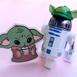 May include: A white and blue R2-D2 droid figurine wearing a green cap with the text YODA printed in yellow, positioned next to a die-cut sticker featuring a green creature with large eyes holding a pink cup with a straw and boba pearls.