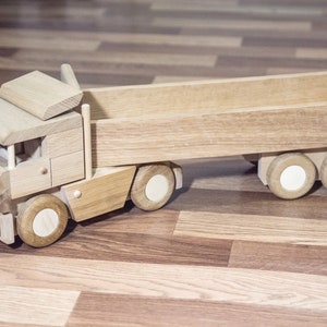 Può includere: Un camion giocattolo in legno con rimorchio, costruito in legno chiaro. Il camion presenta una cabina con tetto, un cassone e sei ruote. Le ruote hanno centri circolari bianchi. Il giocattolo è progettato per i bambini.