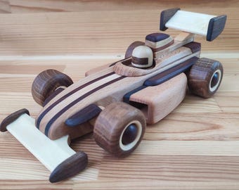 Puede incluir: Un coche de carreras de juguete de madera con carrocería marrón claro, detalles en madera oscura y clara, y una pequeña figura de piloto. El coche tiene ruedas grandes y un alerón trasero.