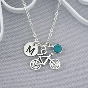 May include: A silver-toned necklace featuring a delicate chain with three charms: a circular pendant engraved with the letter M, a bicycle charm with detailed wheels and frame, and a small, round, teal-colored crystal accent charm.