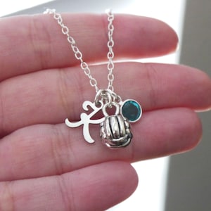May include: A silver-toned chain necklace featuring three charms: a script letter K, a small round blue gemstone, and a detailed silver-toned volleyball charm. The necklace is constructed with a delicate link chain and polished metal components.