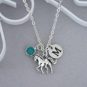 May include: A silver necklace featuring a unicorn charm, a teal-colored gemstone, and a circular pendant with the letter "M". The necklace hangs on a silver chain against a gray background. The unicorn charm is detailed.