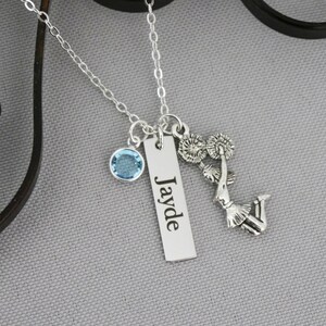 May include: A silver-toned necklace featuring three charms: a round light blue crystal, a rectangular metal bar engraved with the name Jayde, and a detailed silver charm depicting a cheerleader holding pom-poms, all suspended from a delicate chain.