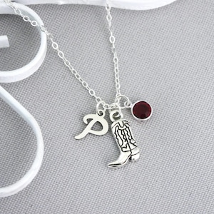 May include: Silver necklace with a charm of a cowboy boot, a letter "P", and a small, round, red gemstone. The necklace is on a gray surface.