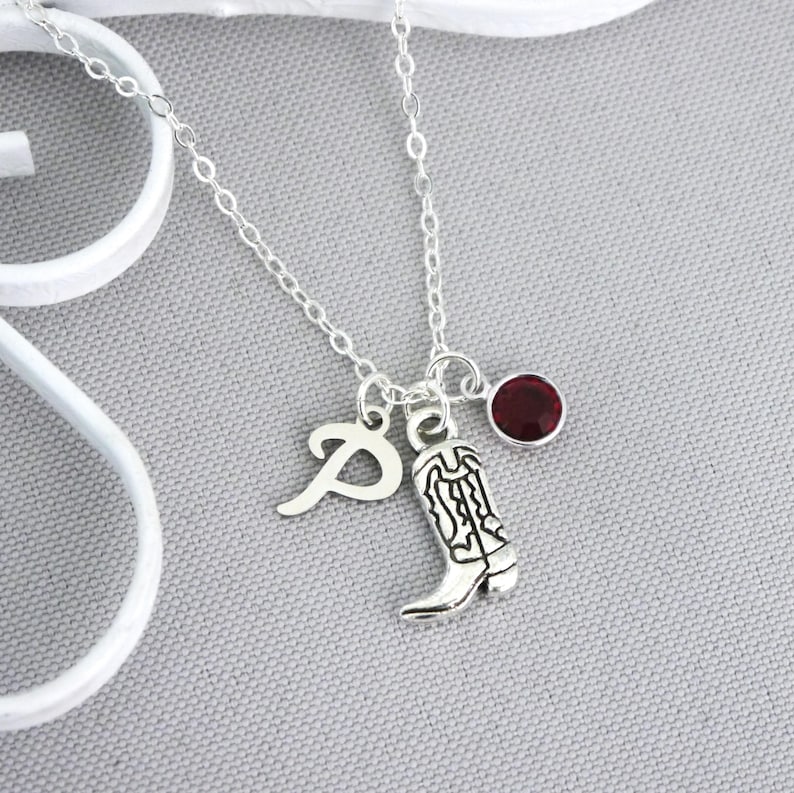 May include: Silver necklace with a charm of a cowboy boot, a letter "P", and a small, round, red gemstone. The necklace is on a gray surface.