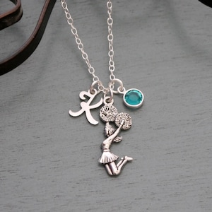 May include: Silver necklace with a cheerleader charm, a letter K charm, and a teal blue gemstone.