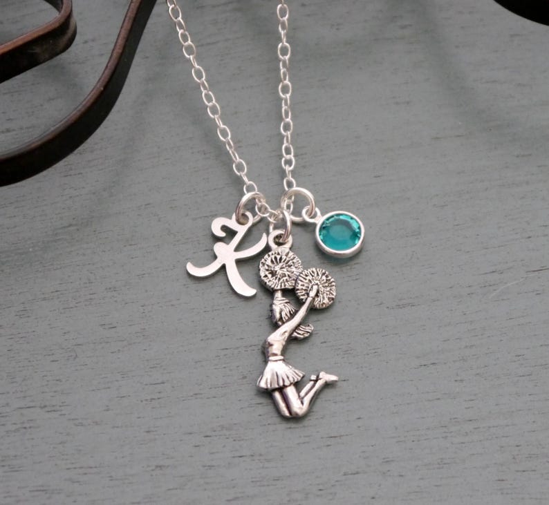 May include: Silver necklace with a cheerleader charm, a letter K charm, and a teal blue gemstone.