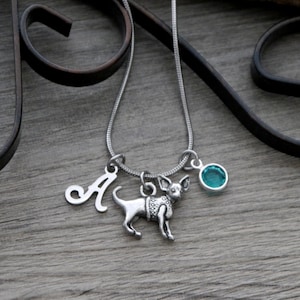 May include: A silver-toned snake chain necklace featuring three charms: a script letter A, a small dog figure with textured detailing, and a round, faceted teal-colored stone set in a silver-toned bezel, all suspended from a single silver-toned ring.