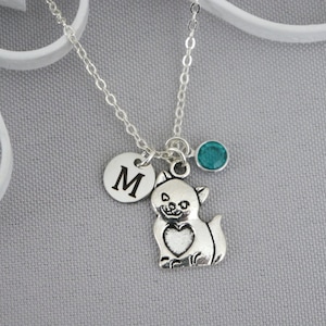 May include: Silver necklace with a small cat charm, a heart, and a round charm with the letter M. A teal blue gemstone is attached to the chain.