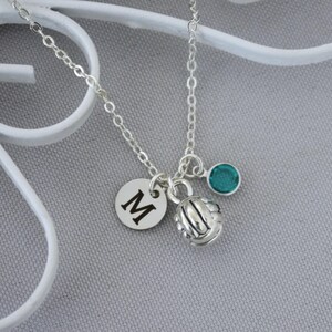 May include: A silver-toned chain necklace featuring three charms: a circular disc engraved with the capital letter M, a three-dimensional volleyball charm with textured panel detailing, and a small round teal-colored crystal bead charm.