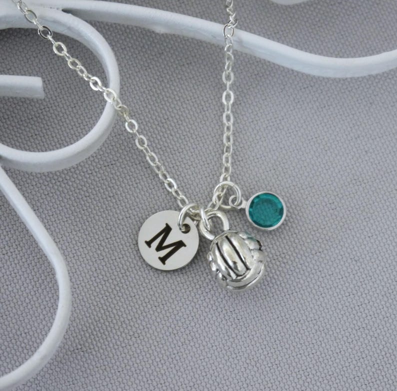 May include: A silver-toned chain necklace featuring three charms: a circular disc engraved with the capital letter M, a three-dimensional volleyball charm with textured panel detailing, and a small round teal-colored crystal bead charm.