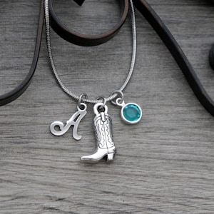 May include: A silver-toned snake chain necklace featuring three charms: a silver letter A, a silver cowboy boot charm with engraved detailing, and a round, bezel-set turquoise-coloured crystal charm hanging from a silver jump ring.