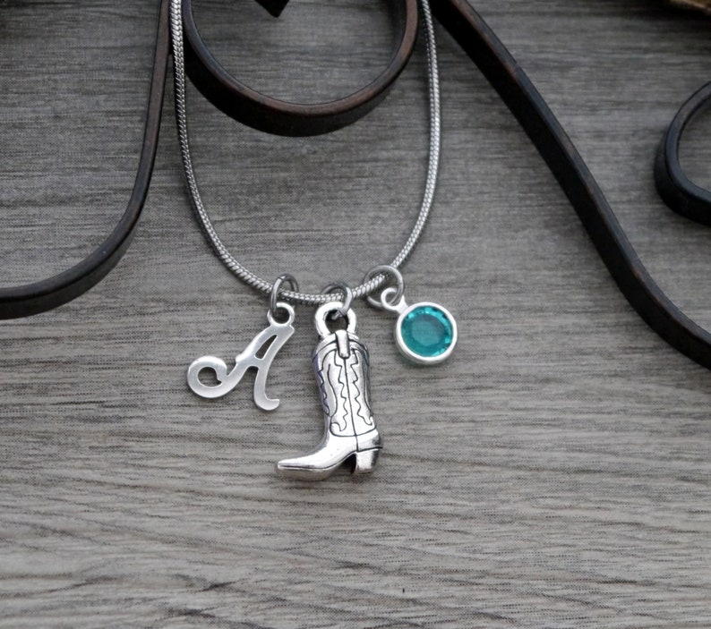 May include: A silver-toned snake chain necklace featuring three charms: a silver letter A, a silver cowboy boot charm with engraved detailing, and a round, bezel-set turquoise-coloured crystal charm hanging from a silver jump ring.